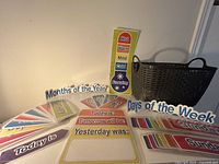 Assorted laminated learning cards for days, months, weather chart and storage basket shown spread on table.