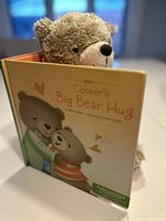 Photo of the children's book 'Cooper's Big Bear Hug' featuring the title and bears hugging on the cover, along with the plush bear peeking from behind the book.