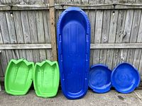 Photo showing 2 green sleds, 1 blue toboggan, and 2 blue saucers lined up against a wooden fence.