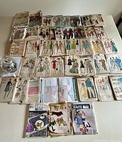 Overall display of assorted vintage sewing patterns laid out on a table showing a wide variety of fashion styles from mid 20th century including dresses, suits, coats for all ages and genders