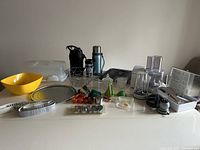 Full view of the kitchenware and small appliances laid out on a surface including food processor, blender, drinkware, fruit basket, mixing bowl, pizza pan, measuring tape, and attachments.