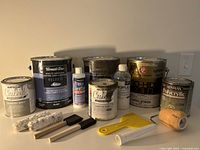 Photo showing various paint cans with different brands and partially used levels, paint rollers, brushes and plastic tools
