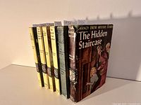 Six Nancy Drew Mystery Stories books standing upright showing front covers, with notable wear on 'The Hidden Staircase' dust jacket, including spine damage and edge wear.