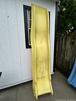 Yellow plastic slide leaning against white building exterior, showing full length and curved sides with some dirt visible.
