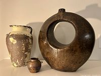 Three rustic decorative vases on white surface with shadow cast. Large circular pottery vase with open center on right, tall distressed pottery vase with handles left, small bronze metal vase centered front.