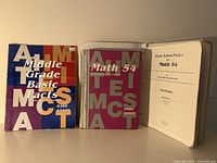 Three Saxon Math 54 educational books displayed side by side including Middle Grade Basic Facts, Math 54 Hard Saxon, and a Home School Packet with test forms