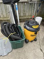 Photo of yellow and black Shop-Vac 14 gallon wet/dry vacuum next to a green Rubbermaid storage bin containing vacuum hoses and accessories, taken outdoors on a concrete surface in front of a wood fence.