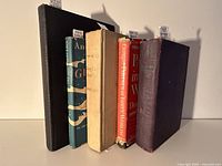 Six vintage hardcover books of varying colors and conditions, shown standing upright against a white background.