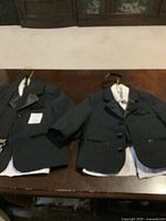 Two black formal suits for small boys hanging on hangers on a wooden table, showing jackets and parts of white shirts underneath.