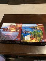 Two 1000-piece Surelox jigsaw puzzles with scenic images placed on a dark wood table, showing both box fronts.