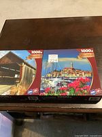 Two Sure-Lox 1000-piece puzzles on a dark wooden table, featuring a covered bridge and a coastal town.