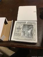 Baptism frame in white styrofoam box, still wrapped in plastic packaging on a wooden table.