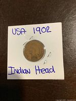 Front side of the 1902 USA Indian Head 1 Cent coin secured in white cardboard holder with handwritten text.