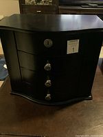 Closed black Bombay mahogany jewelry cabinet with curved front, three drawers, keyhole lock, and silver knobs.