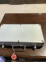 Closed silver aluminum carrying case for poker chips on wooden surface with a red poker chip attached to handle.