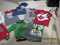 Children's clothing items including several shirts, t-shirts and jackets in assorted colors and designs spread on floor.