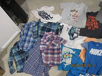 Photo showing assorted children's t-shirts and plaid button-up shirts laid on floor.