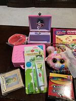 Photo of children's items on table including pink musical jewelry box, Crayola crayons, plush keychain, lip balm/toothbrush set, address book, and activity book.