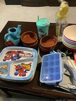 Photo showing variety of children's kitchenware including Spider-Man plates, teapot, brown bowls, ice cube tray, cup with straw, and kitchen towels.