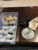 View of plastic container filled with golf balls and assorted wooden and plastic tees, with golf gloves and retractable measuring tape on the side