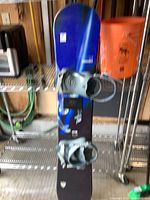 Full view of the Firefly snowboard showing blue and black color with bindings mounted vertically on a metal rack.