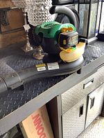 Green and black gas leaf blower with yellow fuel tank cap on a metal surface.