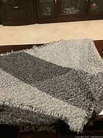 Photo of the ladies grey shawl folded flat on a wooden table, showing the color blocking and fringed edges