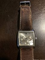 Front view of men's Guess watch showing square silver dial with three subdials and number 12 at top, and worn brown leather strap.