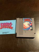 NES game cartridge for Kirby's Adventure front view with instruction booklet beside it on wooden surface.