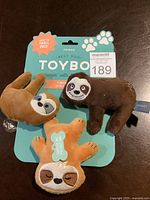 Photo of three small plush dog chew toys shaped like sloths positioned on a blue backing card with 'Best Dog Toy Box' branding and paw prints. Toys have embroidered faces and are designed for small dogs.