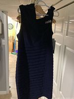 Front view of a dark blue sleeveless dress displaying layered horizontal pleats on the skirt and decorative rhinestone embellishment on one shoulder strap.