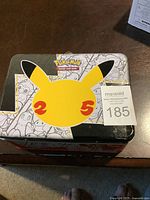 Top view of the Pokémon 25th anniversary lunch box tin featuring a large yellow Pikachu face silhouette with red '25' cheeks and Pokémon Trading Card Game logo on a silver background with black and white Pokémon line art.