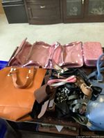 Photo showing multiple purses including orange and blue handbags, assorted belts and straps, and a pink folding makeup bag displayed on carpet.