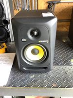 Black KRK Rokit 5 Gen 3 powered studio monitor speaker with yellow woofer cone and black tweeter, front view on metal surface with tools in background.