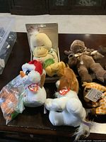 Photo showing white goose plush with red hat, yellow duckling plush in packaging, brown bear plush, and other assorted stuffed animals on a table.