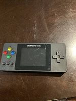 Front view of VX Gaming handheld console with logo visible, directional pad and colored buttons on sides, screen off.