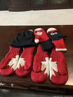 Wide view of three pairs of mittens and one toque hat showing colors and maple leaf designs.