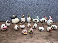 Lot of 17 handmade porcelain eggs featuring various bird figurines emerging from cracked eggshells, all arranged on wood surface