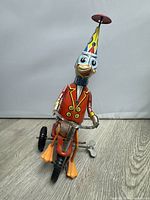 Front view of the colorful 1950s wind-up tin toy duck wearing a party hat and sitting on a tricycle, showing the missing rear wheel on the left side.