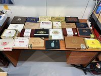 Photo of 24 wooden cigar boxes arranged on a brown desk, showing various brands and designs including Montecristo, Punch, New World, and others.