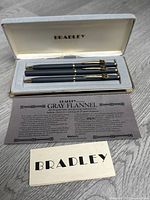 Set of three writing instruments in original white presentation box with 'BRADLEY' inscription on inside lid. Items have gray flannel bodies with gold plated clip and trim. Includes original documentation card explaining Gray Flannel materials and quality.