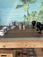 Wide shot of the lot displaying various drinking glasses and mugs arranged on a wooden table against a tropical wall mural.