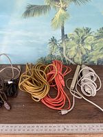 Photo showing multiple extension cords of various colors and thicknesses coiled on a wooden surface in front of a tropical-themed backdrop. Includes orange, yellow, red heavy-duty cords and smaller brown and beige cords.