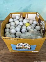 Box half full of used white and a few yellow golf balls of mixed brands in a cardboard box labeled Newman Organics and Keurig Hot.