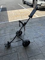 Side view of black pull behind golf cart with three wheels and adjustable handle, standing on pavement outdoors.
