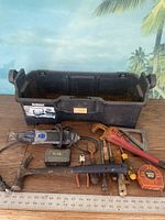 Various handyman tools including Dremel Multi-Max MM30 oscillating power tool, claw hammer, pipe wrench, tape measure and other metal hand tools arranged on wooden surface