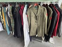 Wide view of hanging garments on rack showing two military uniform jackets with red insignia patches and various other clothing items including jackets, dresses, and sweatshirts on mostly plastic hangers.