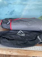 View of folded Hillary tent in gray and red bag next to black bag with Hillary logo laid on a wooden surface.