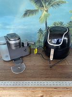 Pair of kitchen appliances on wooden surface with a tropical background; left is Keurig coffee maker and right is Power XL air fryer, both powered with cords attached.
