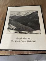Framed black and white photographic print of a mountain landscape titled 'Ansel Adams - The Mural Project 1941-1942' placed on a wooden surface.